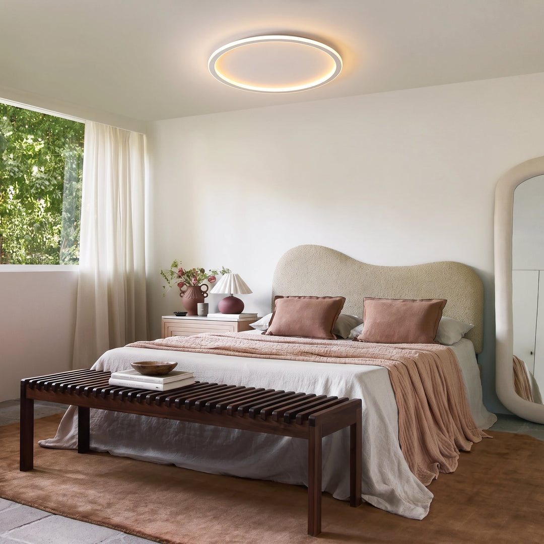 Plafonnier annulaire allumé dans une chambre aux tons rosés, avec lit à tête arrondie, banc en bois et grand miroir. 11