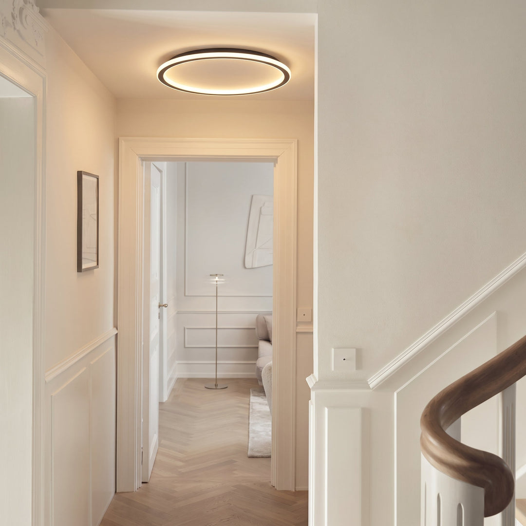 Plafonnier en anneau allumé dans un couloir blanc à moulures, avec un escalier à rampe en bois et une porte ouverte. 5