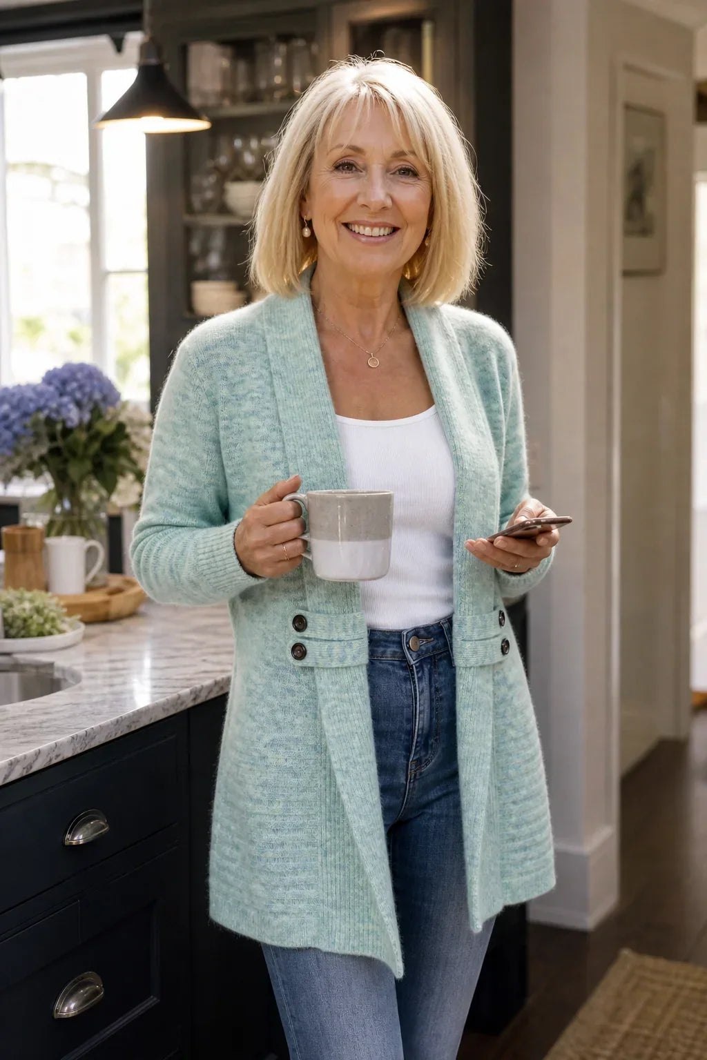 Femme blonde souriante en cardigan vert d'eau, tasse et téléphone en main, dans une cuisine 4