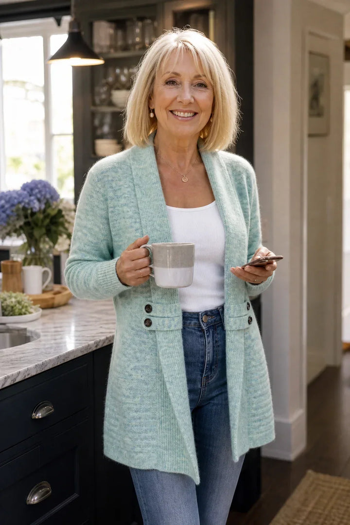 Femme blonde souriante en cardigan vert d'eau, tasse et téléphone en main, dans une cuisine 4