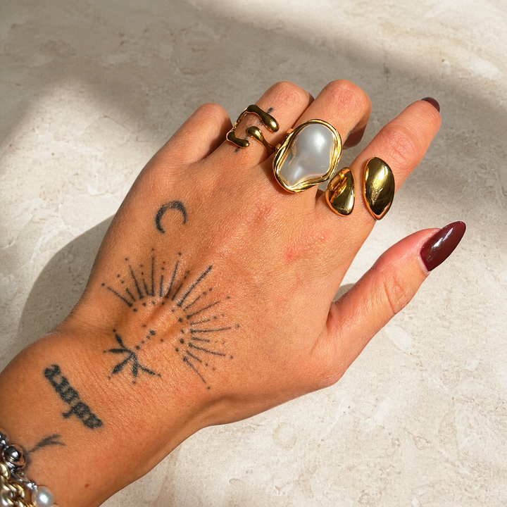 Main tatouée posée sur une surface en pierre claire, portant la bague dorée ouverte avec d'autres bagues, dont une à grande perle. 4