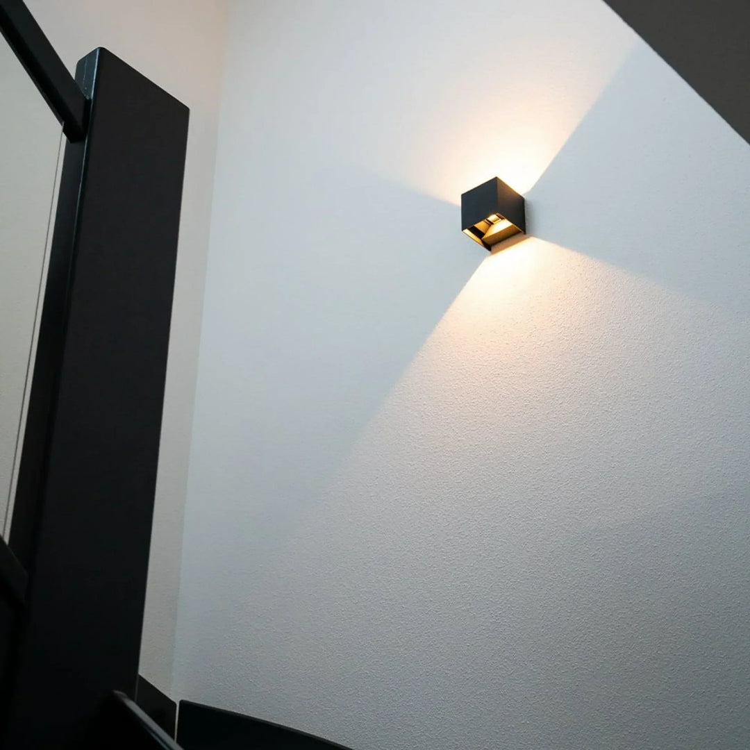 Applique cubique noire allumée sur un mur blanc de cage d'escalier, vue en contre-plongée près d'une rampe noire 8