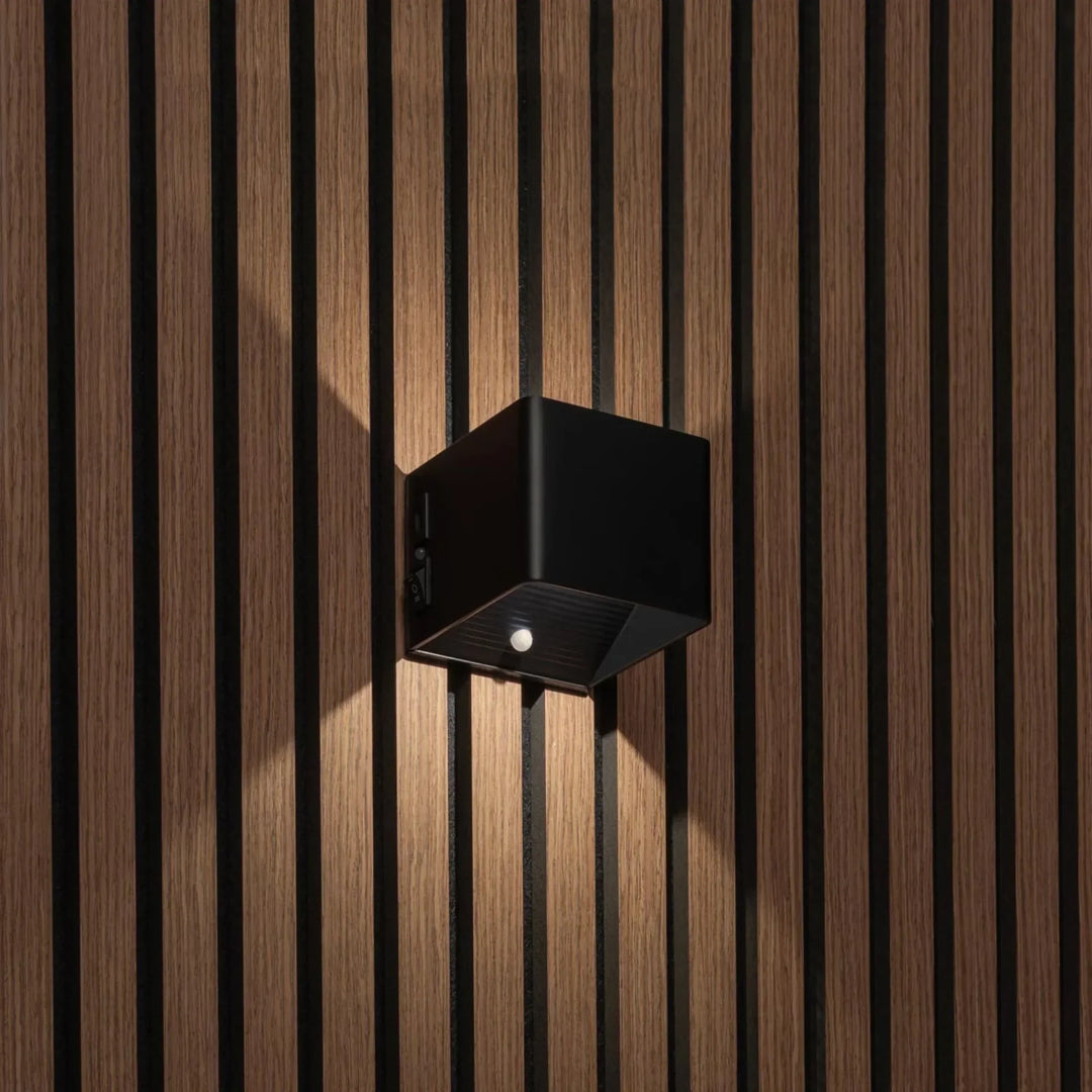 Applique cubique noire allumée sur un mur à lattes de bois, vue de trois quarts montrant le détecteur et l'interrupteur 1