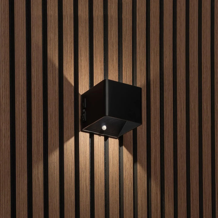 Applique cubique noire allumée sur un mur à lattes de bois, vue de trois quarts montrant le détecteur et l'interrupteur 1