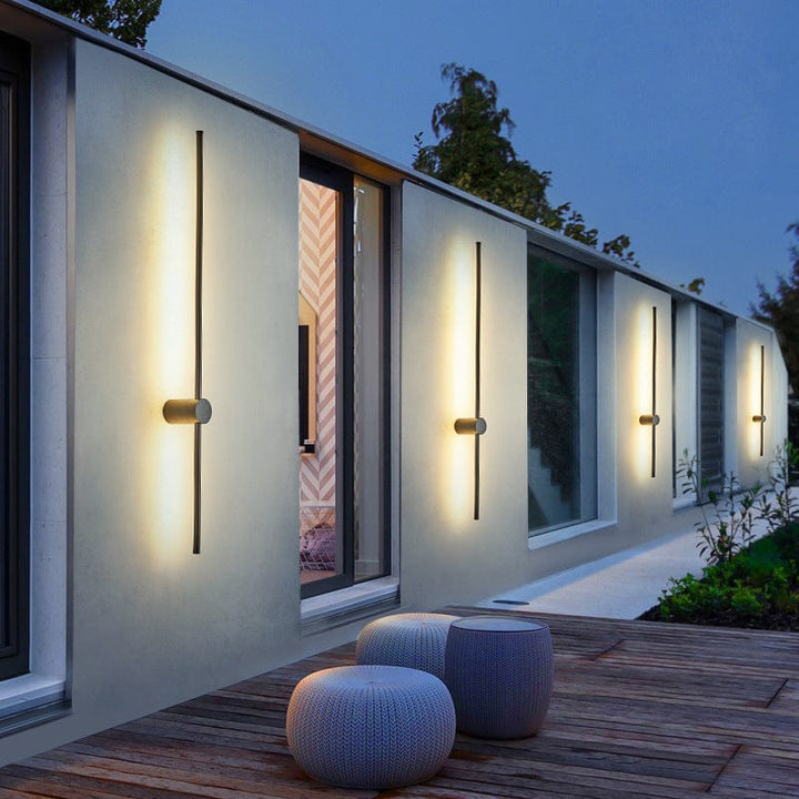 Quatre appliques fines noires alignées sur la façade claire d'une maison au crépuscule, avec des poufs en tricot sur la terrasse en bois. 1