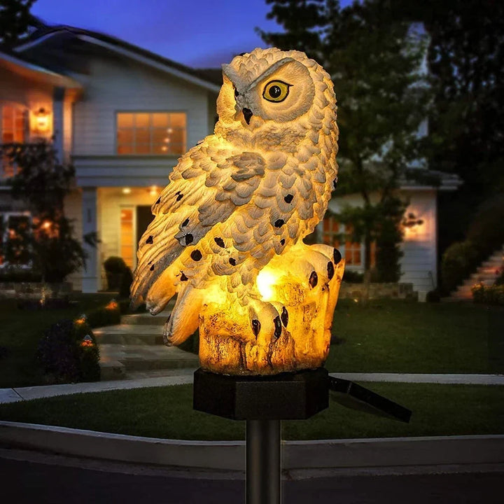 Hibou blanc moucheté de noir éclairé de l'intérieur, planté sur son pieu dans un jardin devant une maison illuminée le soir. 2