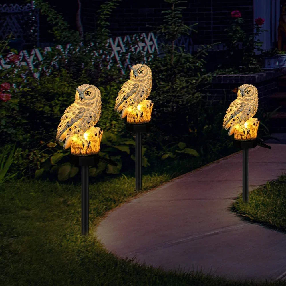Trois hiboux lumineux alignés au bord d'une allée de jardin, de nuit, parmi les plantes et les fleurs. 3