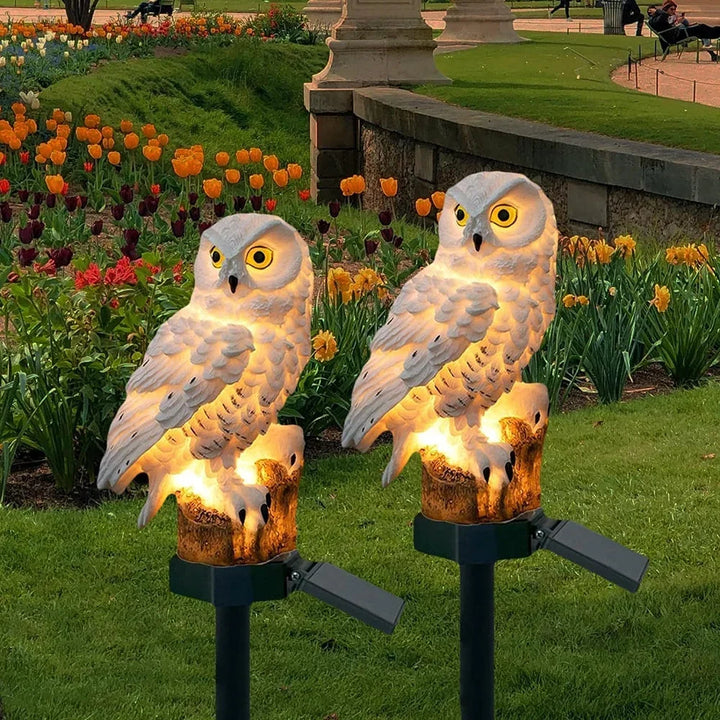 Deux hiboux blancs lumineux plantés dans une pelouse au crépuscule, devant des parterres de tulipes orange et de jonquilles. 1