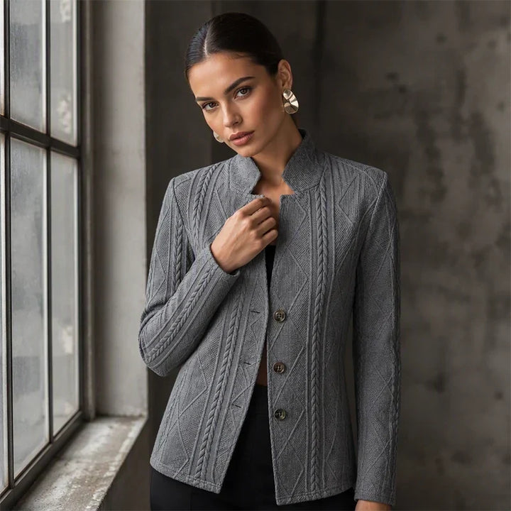Femme posant près d'une fenêtre, la main sur le col montant du blazer gris foncé aux motifs de torsades et losanges. 10