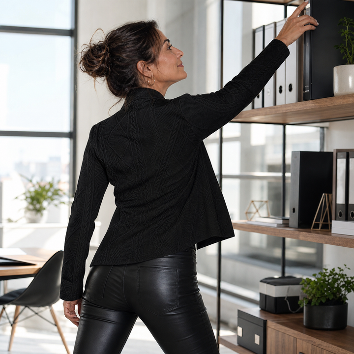 Vue de dos d'une femme au bureau, bras levé vers une étagère, portant le blazer noir texturé et un pantalon en cuir noir. 13