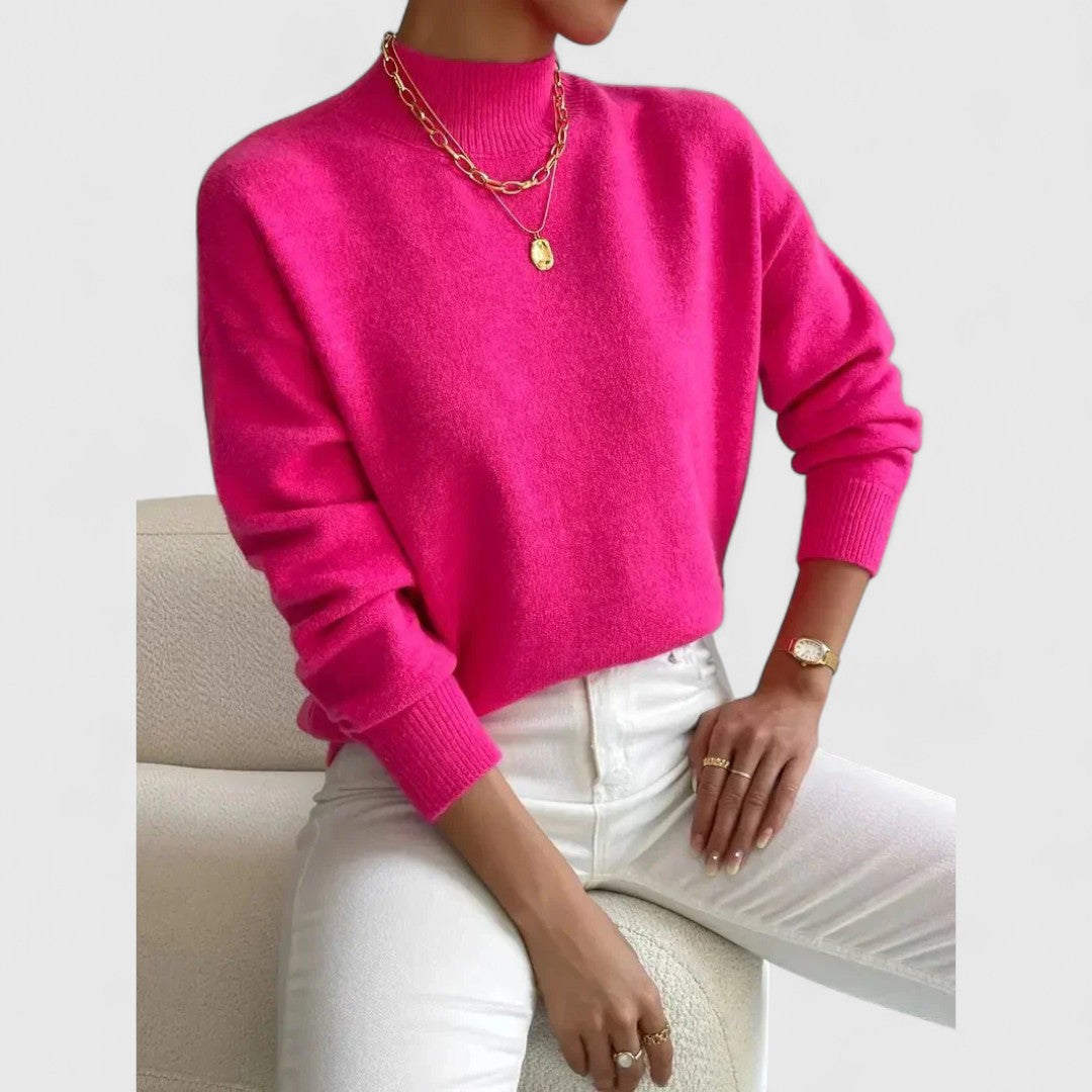 Pull rose fuchsia vif à col montant porté par une femme assise, avec pantalon blanc et colliers dorés. 9