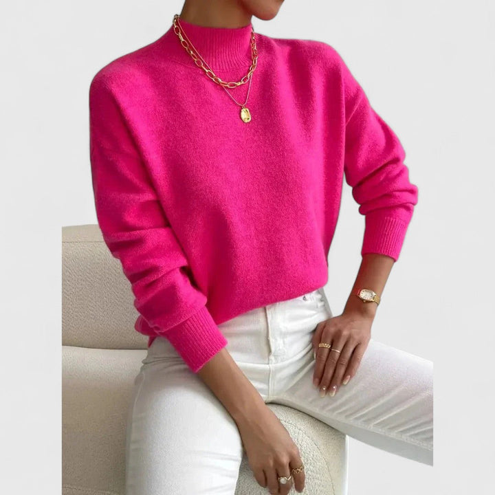 Pull rose fuchsia vif à col montant porté par une femme assise, avec pantalon blanc et colliers dorés. 9