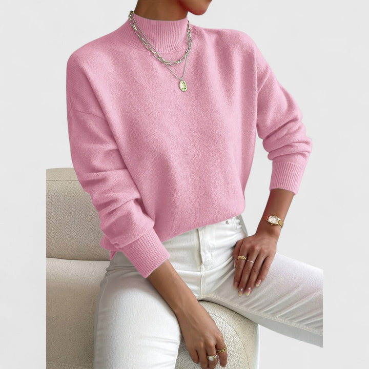 Pull rose bonbon à col montant et poignets côtelés, porté avec un pantalon blanc, assise sur un fauteuil bouclé. 5