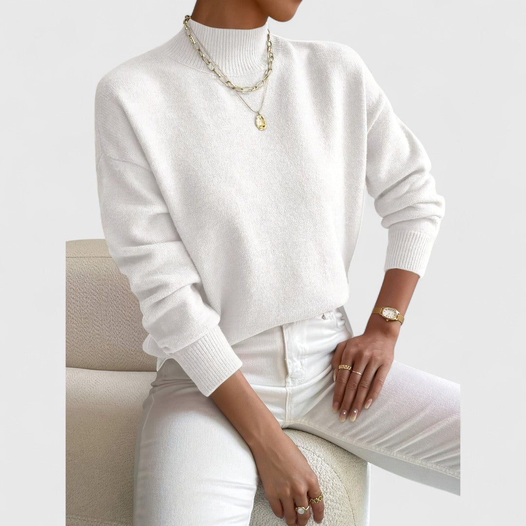 Pull blanc crème à col montant porté par une femme assise, avec pantalon blanc, collier doré et montre. 4