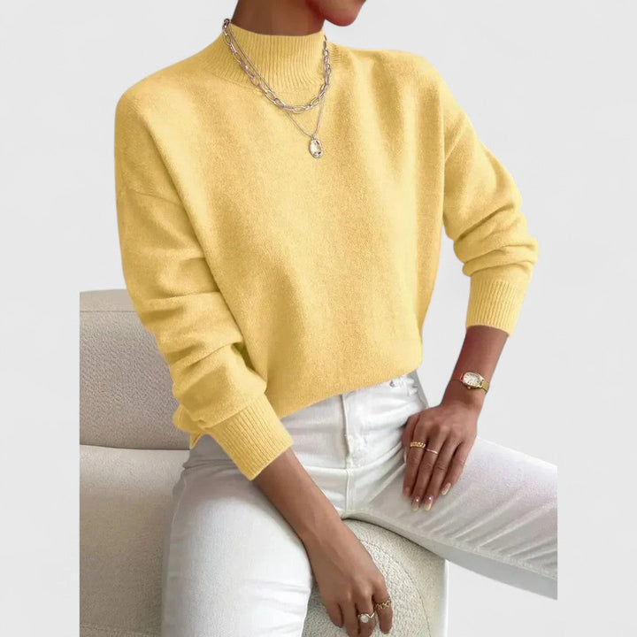 Pull jaune pastel à col montant porté par une femme assise, avec pantalon blanc et collier argenté à pendentif. 6
