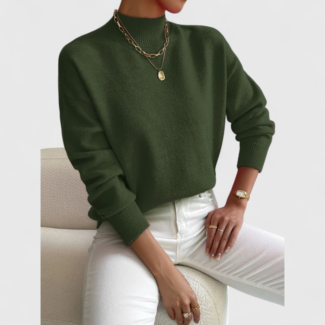 Pull vert kaki foncé à col montant, porté avec un pantalon blanc, collier doré et bagues, sur fauteuil crème. 10