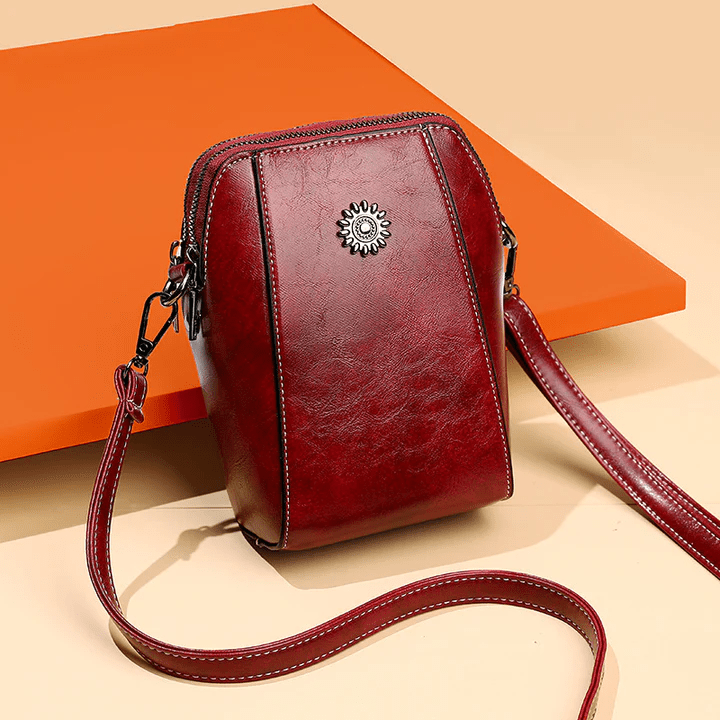 Sac en cuir rouge bordeaux avec ornement fleur, posé contre un panneau orange, sangle étalée 7