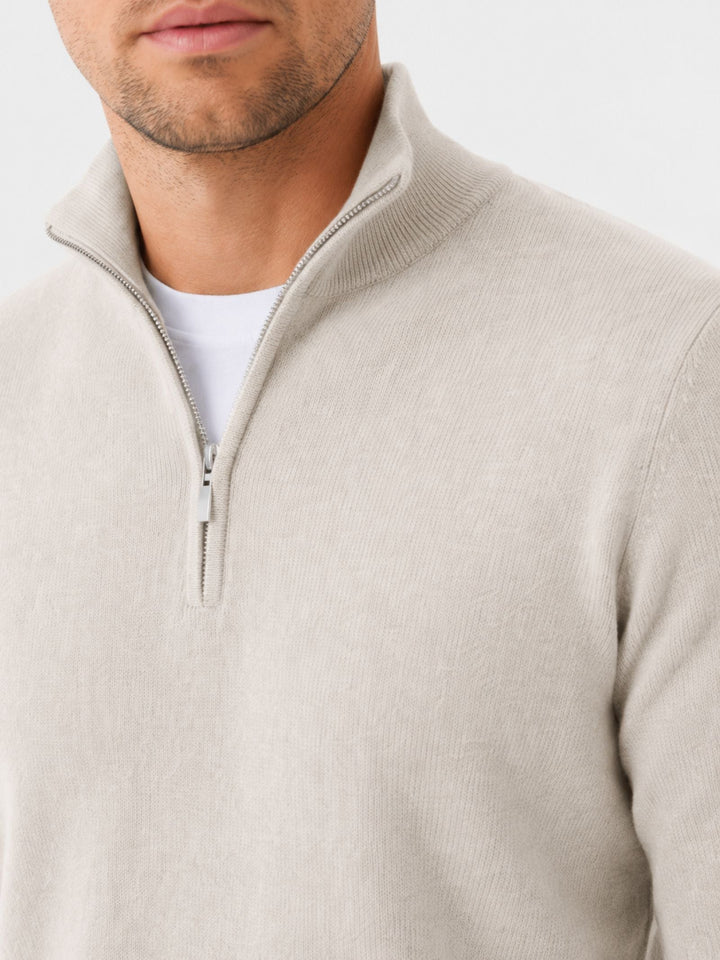 Gros plan sur le col montant et la fermeture éclair métallique du pull beige porté par un homme. 11