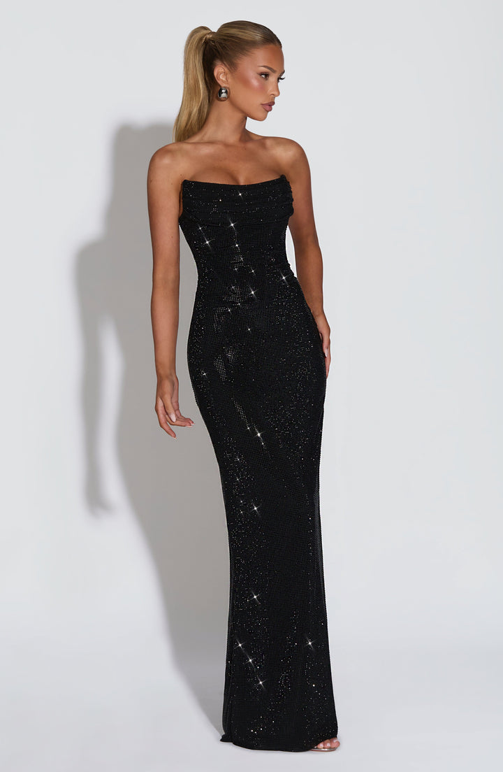 Robe longue noire à strass portée de trois quarts, buste drapé, coupe ajustée jusqu'aux chevilles et sandales argentées visibles. 25
