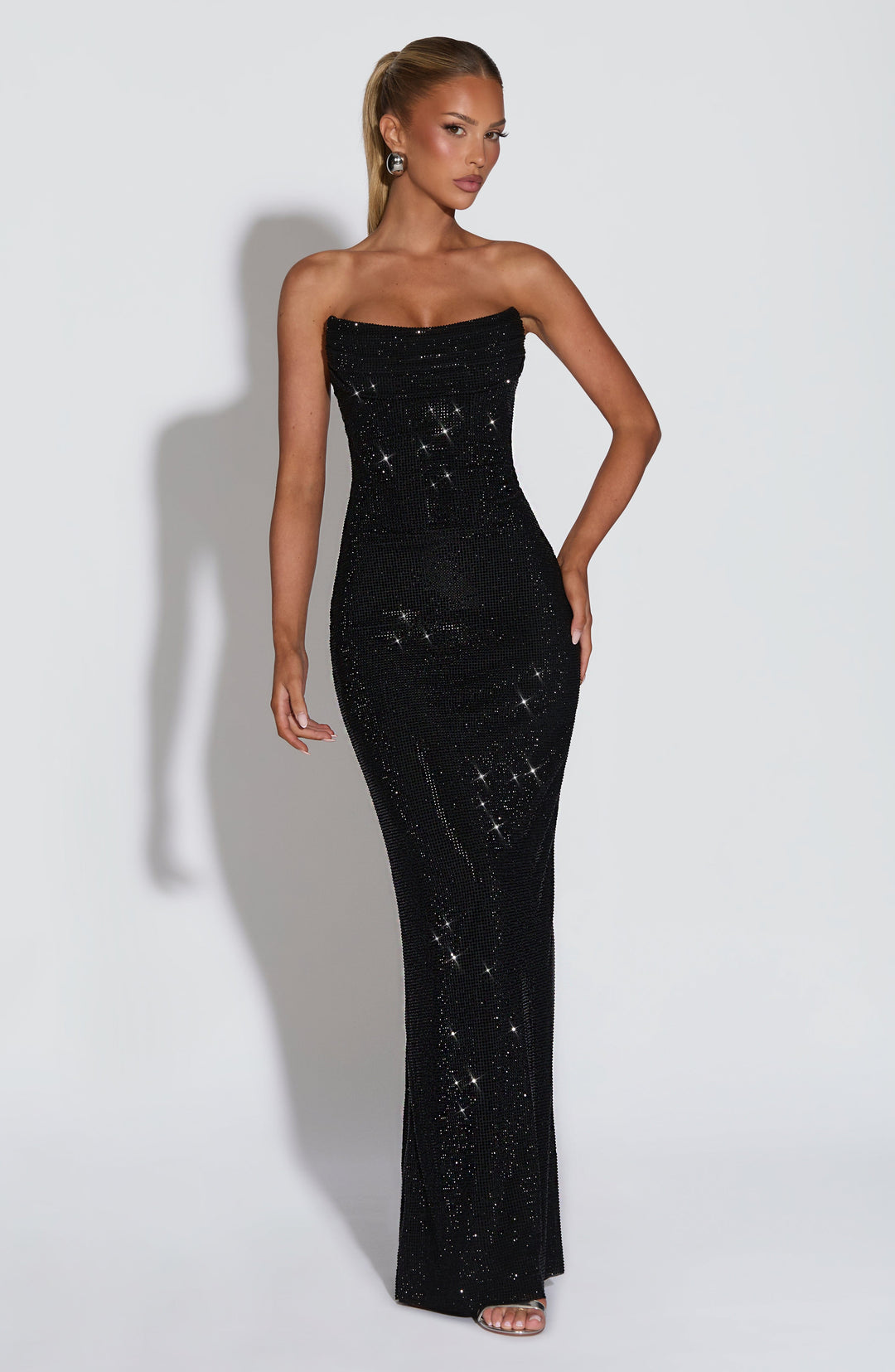 Robe noire à strass portée de face par un mannequin à queue-de-cheval haute, une main sur la hanche, silhouette fourreau jusqu'au sol. 22
