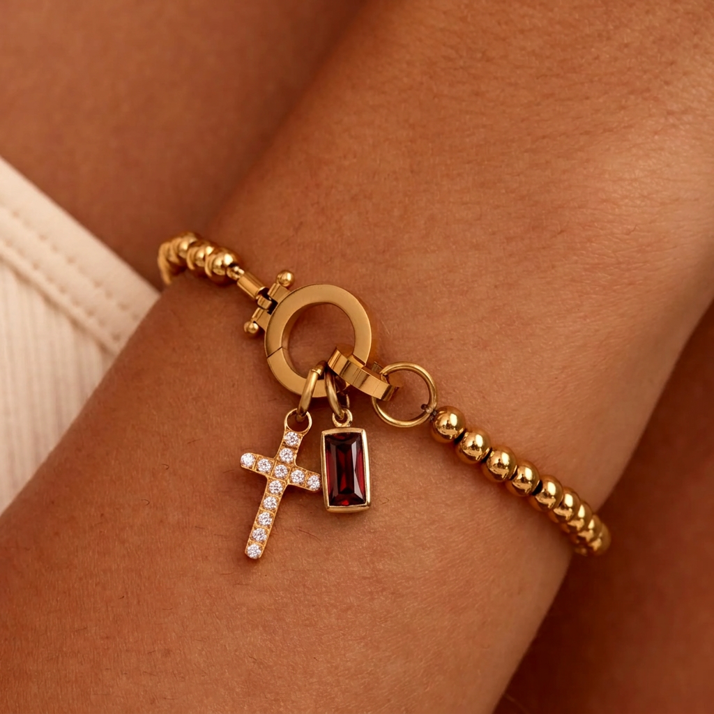 Bracelet doré à perles porté au poignet, avec croix sertie de strass et pierre rouge foncé rectangulaire. 15