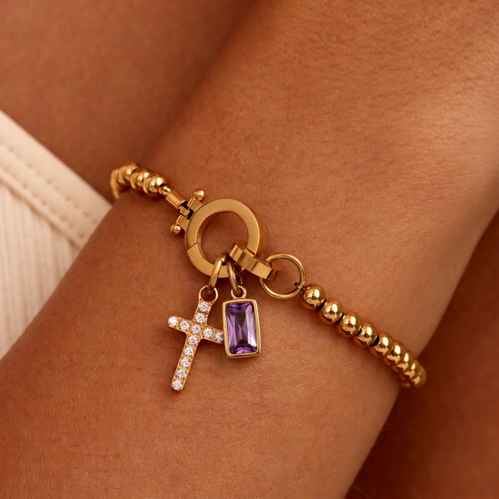 Bracelet doré à perles porté au poignet, avec croix sertie de strass et pierre violette rectangulaire. 17