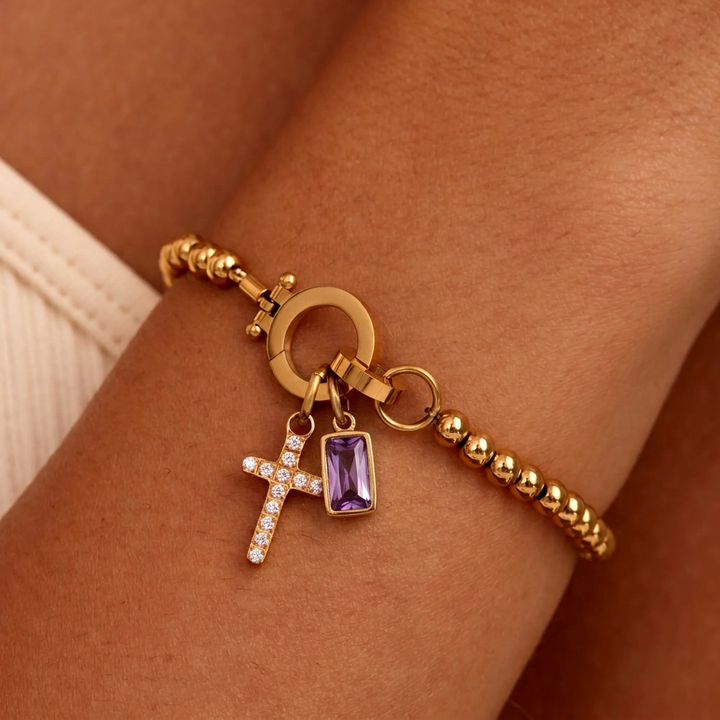 Bracelet doré à perles porté au poignet, avec croix sertie de strass et pierre violette rectangulaire. 17