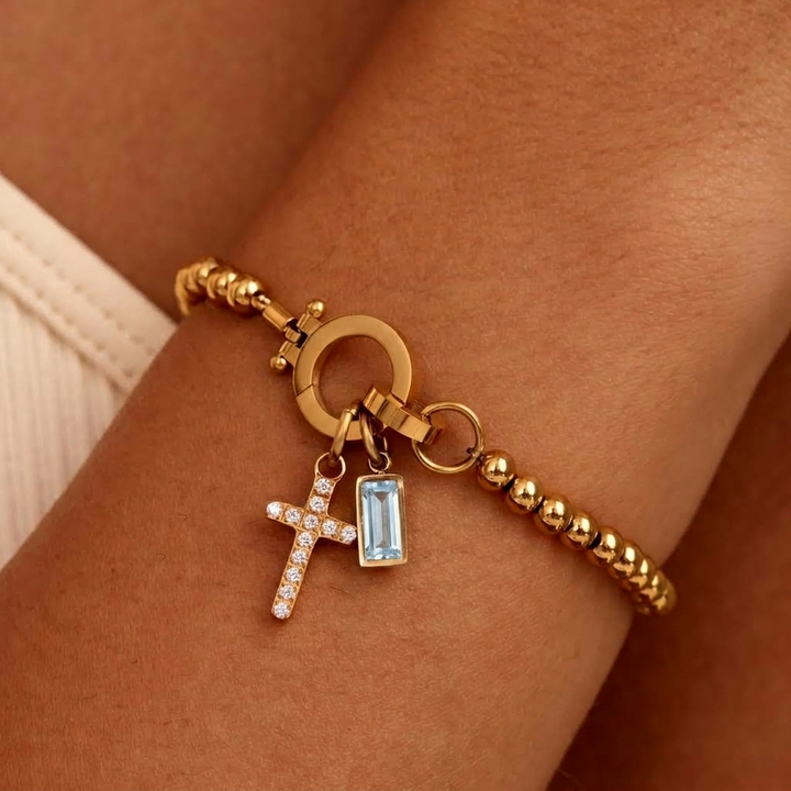 Bracelet doré à perles porté au poignet, avec croix sertie de strass et pierre bleu clair rectangulaire. 19