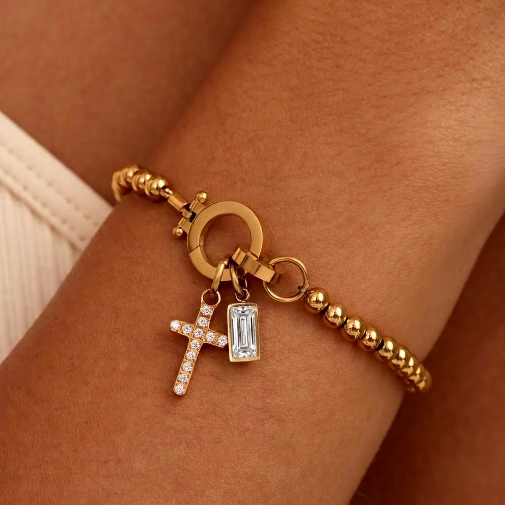 Bracelet de petites billes dorées porté au poignet, avec breloque croix pavée de strass et pierre transparente rectangulaire. 21