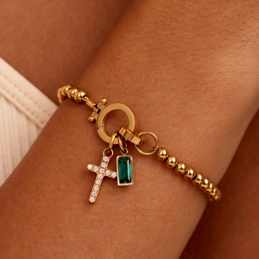 Bracelet de billes dorées porté au poignet, avec breloque croix pavée de strass et pierre rectangulaire vert émeraude. 23