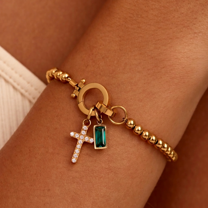 Bracelet de billes dorées porté au poignet, avec breloque croix pavée de strass et pierre rectangulaire vert émeraude. 23