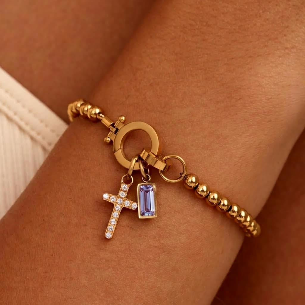 Bracelet de billes dorées porté au poignet, avec breloque croix pavée de strass et pierre rectangulaire violet clair. 25