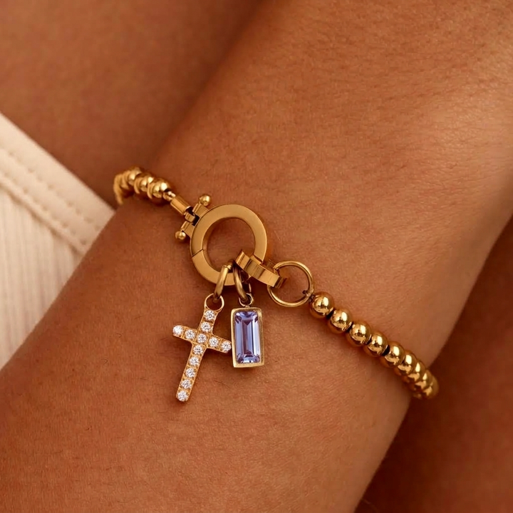 Bracelet de billes dorées porté au poignet, avec breloque croix pavée de strass et pierre rectangulaire violet clair. 25