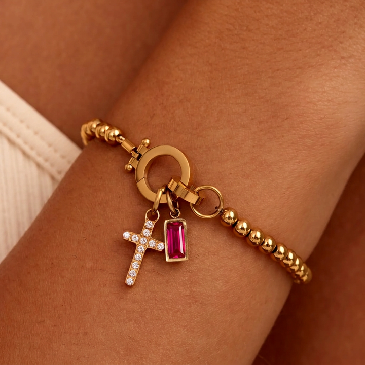Bracelet de billes dorées porté au poignet, avec breloque croix pavée de strass et pierre rectangulaire rose fuchsia. 27