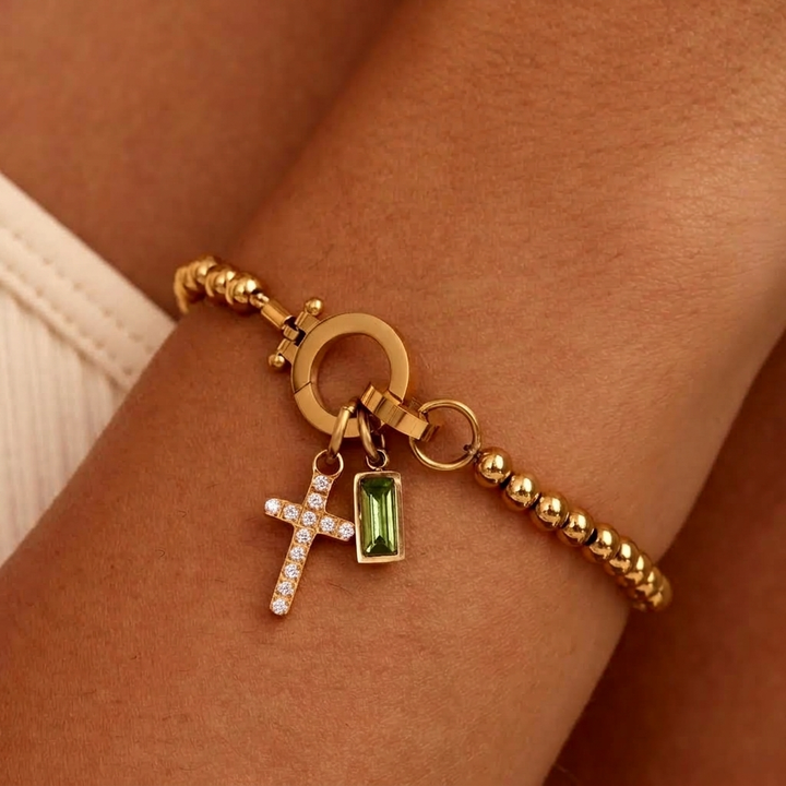 Bracelet de billes dorées porté au poignet, avec breloque croix pavée de strass et pierre rectangulaire vert clair. 29