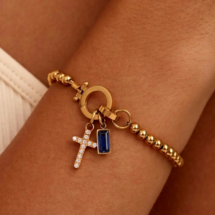Bracelet de billes dorées porté au poignet, avec breloque croix pavée de strass et pierre rectangulaire bleu saphir. 31