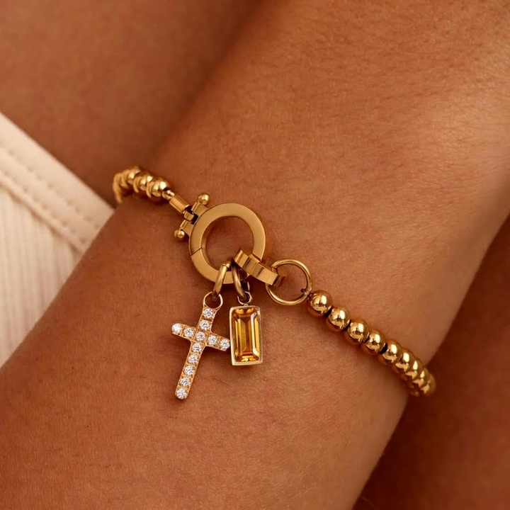 Bracelet de billes dorées porté au poignet, avec breloque croix pavée de strass et pierre rectangulaire jaune ambré. 35