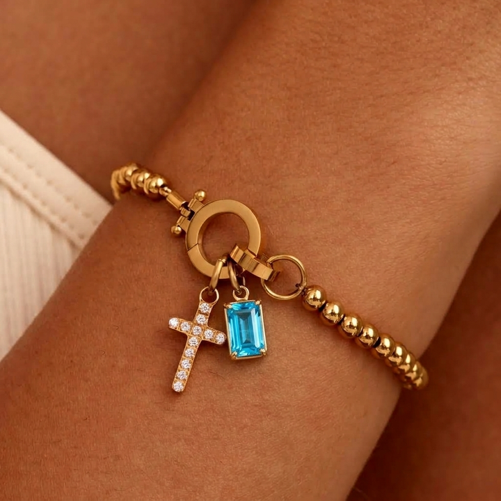 Bracelet de billes dorées porté au poignet, avec breloque croix pavée de strass et pierre bleu turquoise sertie de griffes. 37
