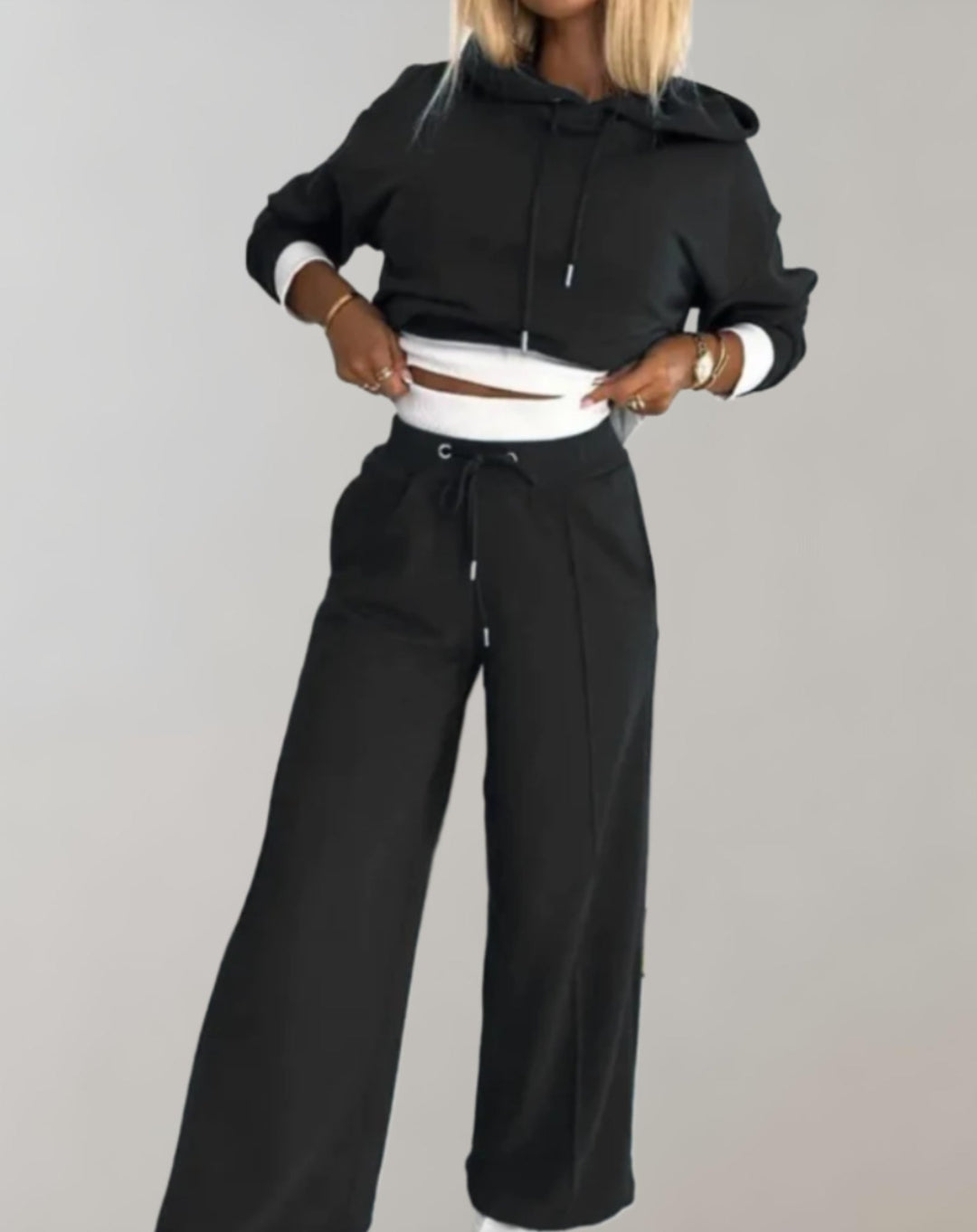 Femme portant l'ensemble noir, sweat court à capuche aux poignets blancs relevé sur la taille et pantalon large à cordon. 1