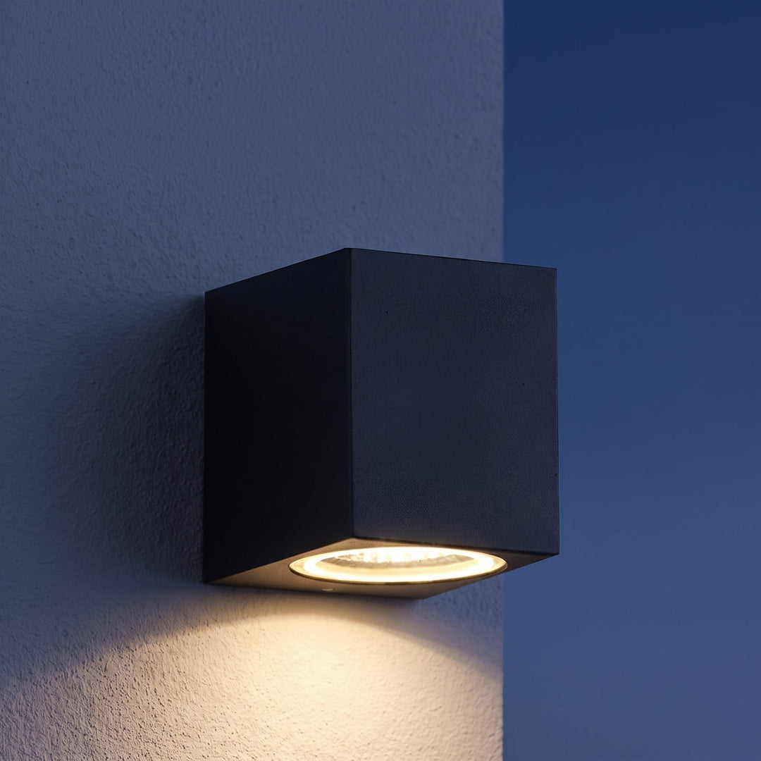 Applique cubique noire fixée à un mur texturé, diffusant une lumière chaude vers le bas au crépuscule 2