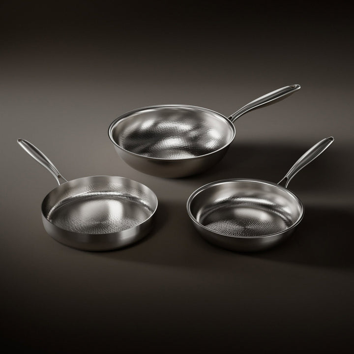 Trois poêles à surface martelée brillante, un wok et deux poêles à manche en acier, posées sur un fond sombre. 1