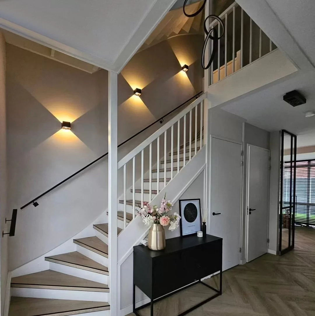 Trois appliques cubiques noires alignées le long d'un escalier blanc, éclairant le mur dans un hall avec console noire et fleurs. 7