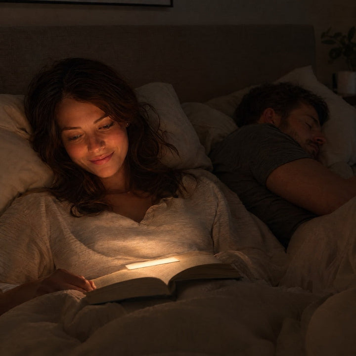 Femme souriante lisant dans son lit à la lumière douce posée sur le livre, son conjoint dormant 2