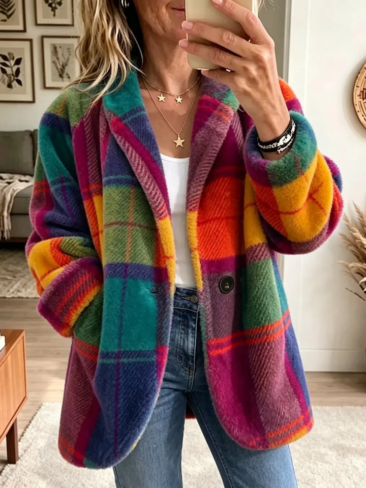 Femme portant le manteau moelleux à grands carreaux orange, vert, jaune et violet, ouvert sur un haut blanc et un jean, dans un salon. 1