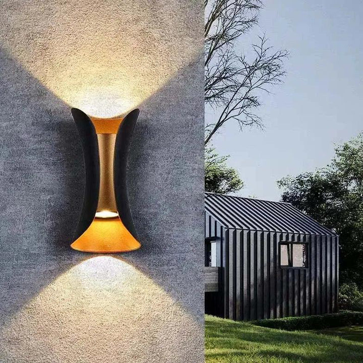 Gros plan de l'applique noire et dorée sur un mur gris texturé, projetant un faisceau lumineux vers le haut et vers le bas, avec une maison sombre à côté. 1