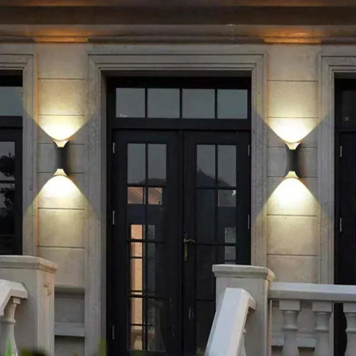 Deux appliques noires allumées de part et d'autre d'une porte-fenêtre noire, sur une façade de pierre claire, éclairant le mur vers le haut et le bas. 2