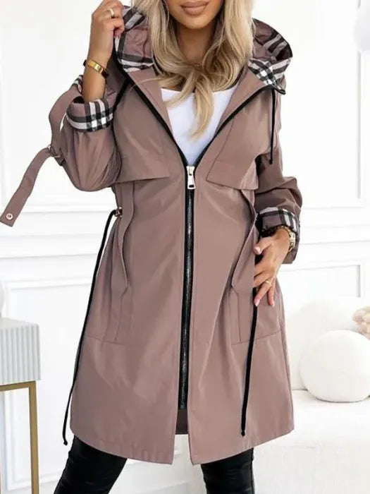 Veste mi-longue taupe portée par une mannequin, capuche et poignets doublés à carreaux, taille ajustée par cordons, zip ouvert sur un haut blanc. 1