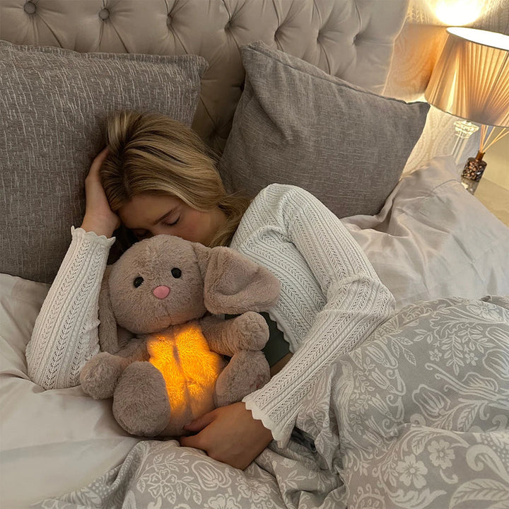 Femme blonde endormie, tenant contre elle le lapin en peluche dont le ventre brille, entre coussins gris et lampe. 16