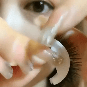 Gros plan sur une main qui décolle de la paupière un support blanc en arc portant des cils, près d'un œil. 14