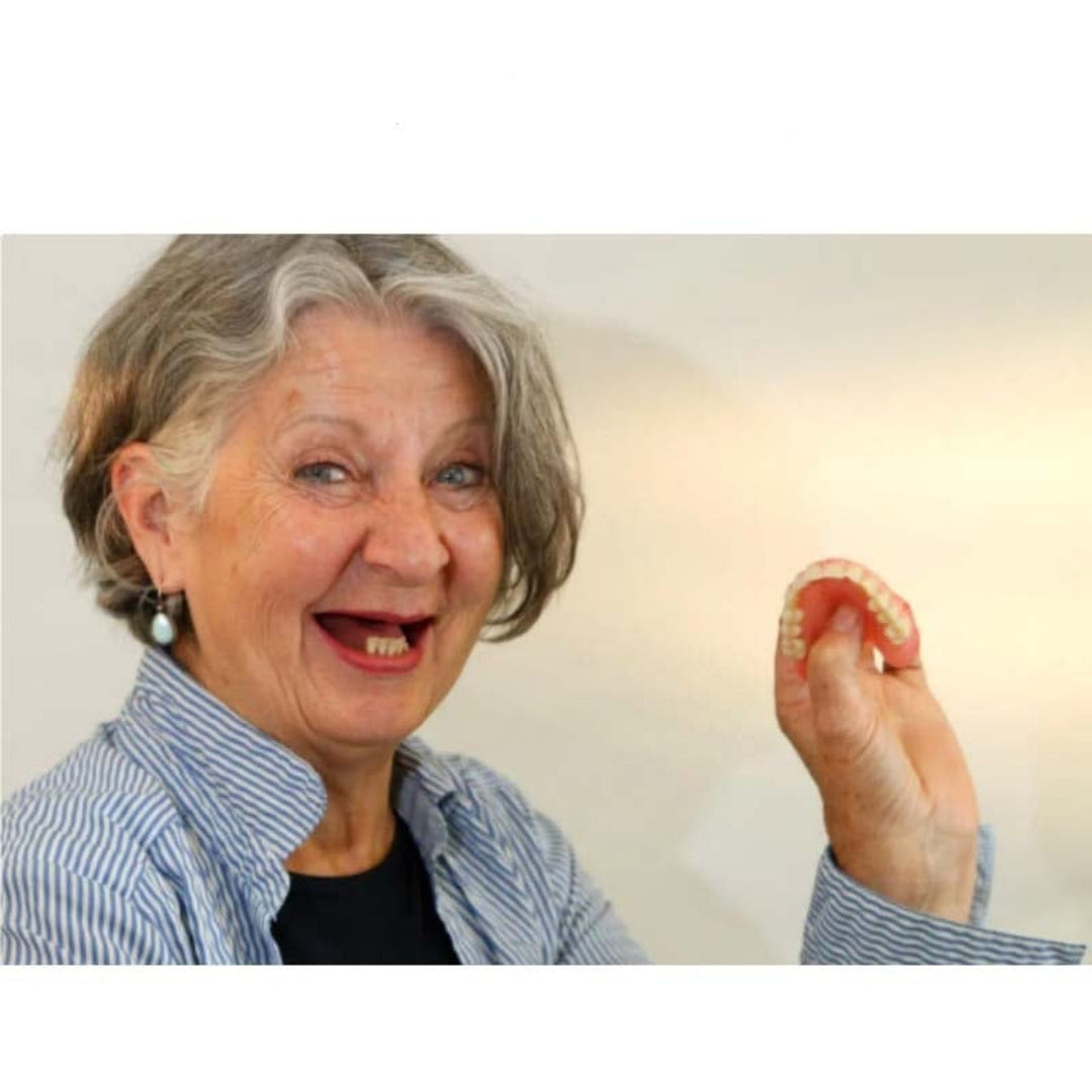 Femme aux cheveux gris souriante, dents du haut absentes, qui tient et plie entre ses doigts une prothèse dentaire souple. 3